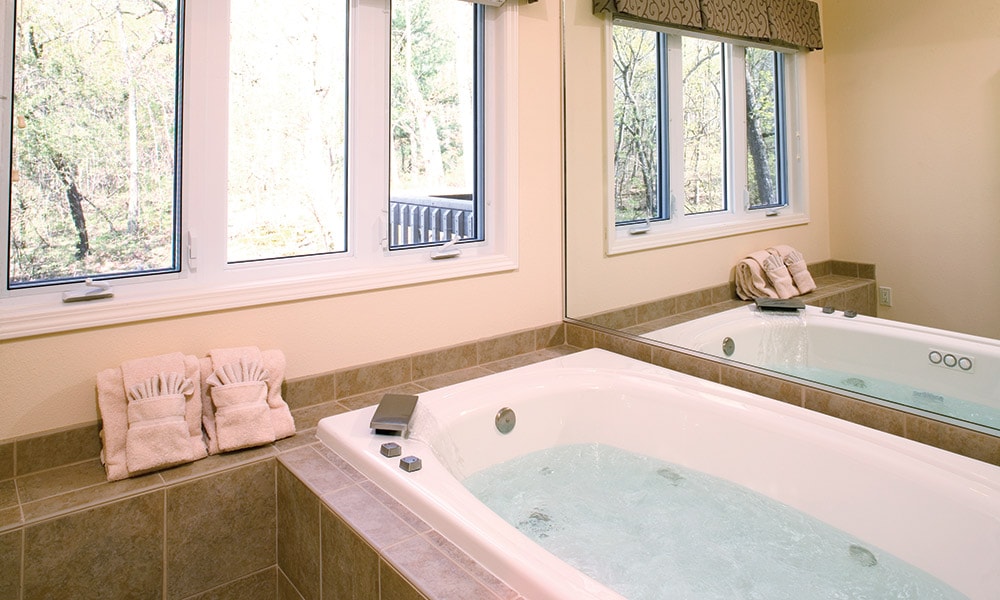 A spacious bathroom features a relaxing jetted tub positioned beside large windows, allowing natural light to enter. Two plush towels are neatly arranged on the counter, complementing the clean lines of the neutral tile and serene ambiance created by the surrounding natural scenery.