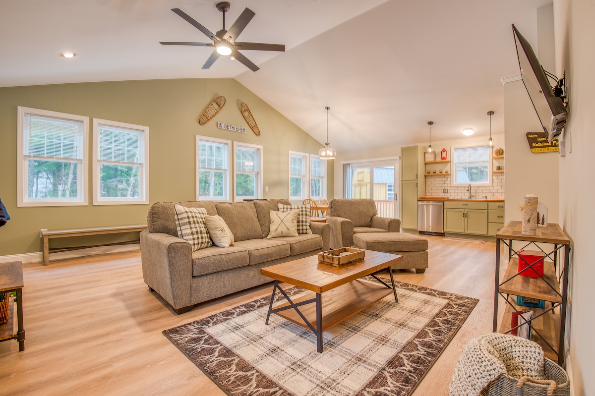 A spacious living area is highlighted by large windows allowing natural light. Comfortable seating is arranged around a coffee table, while a light-colored area rug adds warmth. Modern decor includes a flat-screen TV mounted on the wall, and a kitchen area is visible in the background.