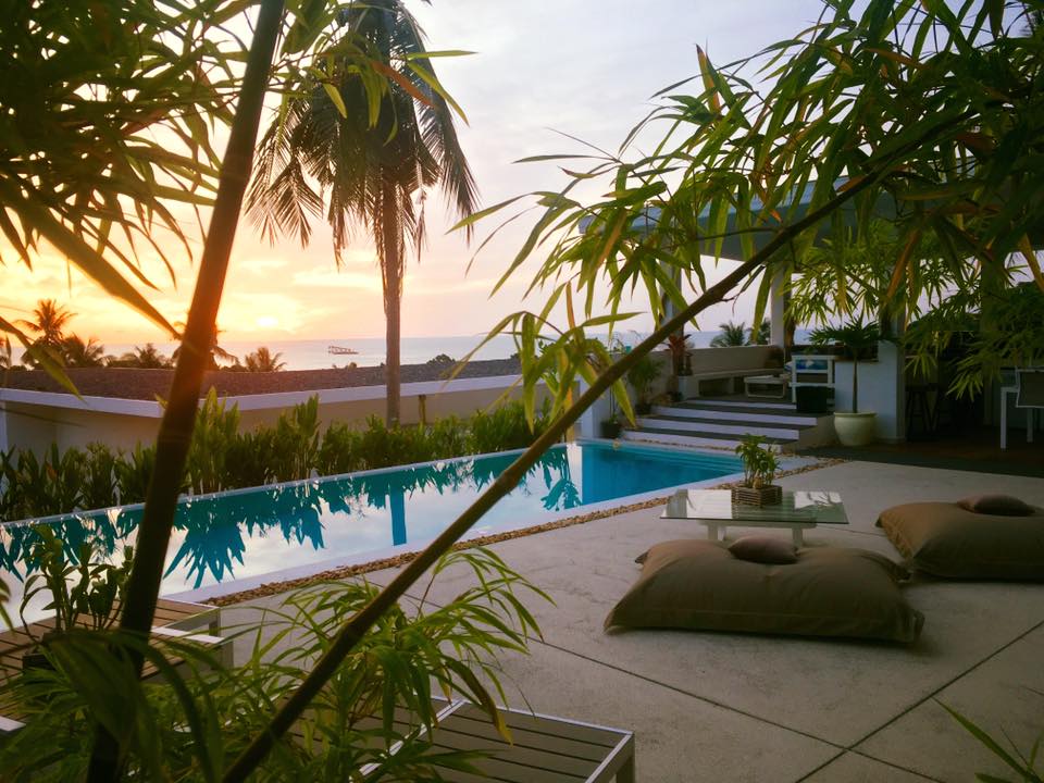 The image captures a serene outdoor space featuring a shimmering infinity pool surrounded by lush green foliage. Comfortable seating lies beside the pool, and a warm sunset casts a golden glow over the landscape, enhancing the tranquil atmosphere.