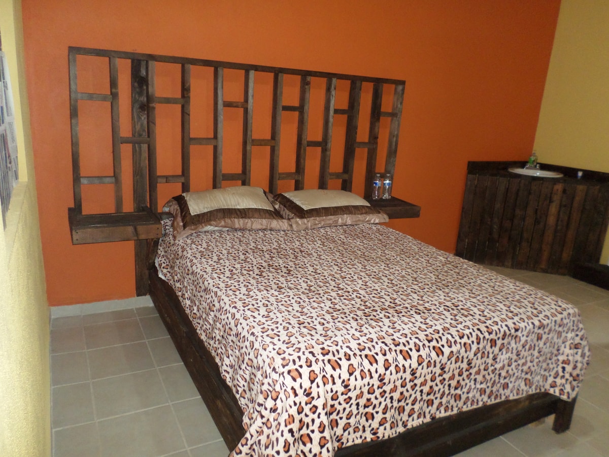 A spacious bed with a leopard print coverlet sits against a vibrant orange wall. The headboard of rustic wood features an open geometric design. Two bottles of water are placed on the bedside ledge, enhancing the room's functionality.