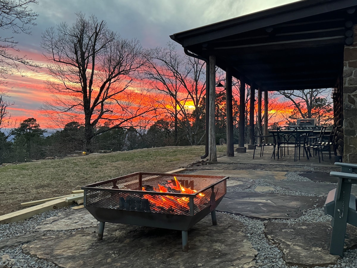 A fire pit sits on a stone patio, with flames glowing warmly. Beyond, a vibrant sunset paints the sky in shades of orange and pink, framed by silhouetted trees. A dining table with chairs is visible under an overhanging roof, creating an inviting outdoor space.
