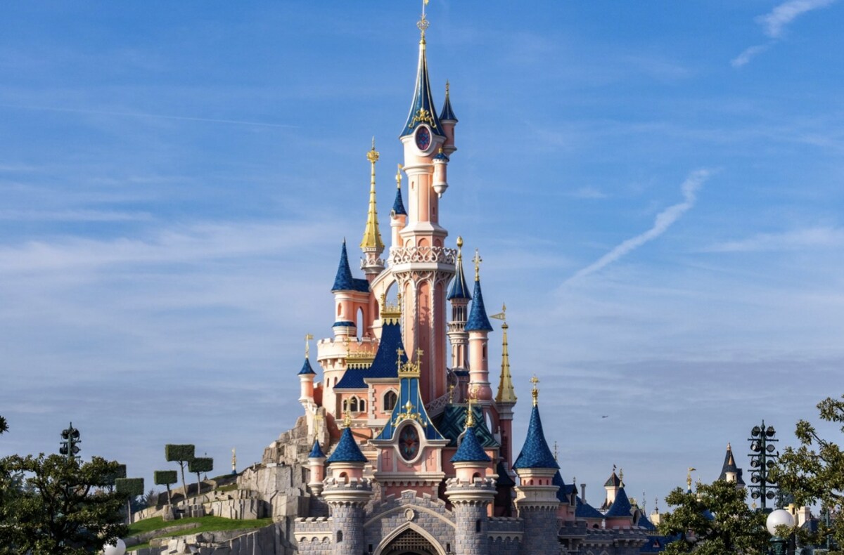 A whimsical castle structure towers against a clear blue sky. The exterior features pink and blue hues, along with pointed turrets and ornate detailing. Lush greenery surrounds the castle, adding to the enchanting ambiance of the scene.