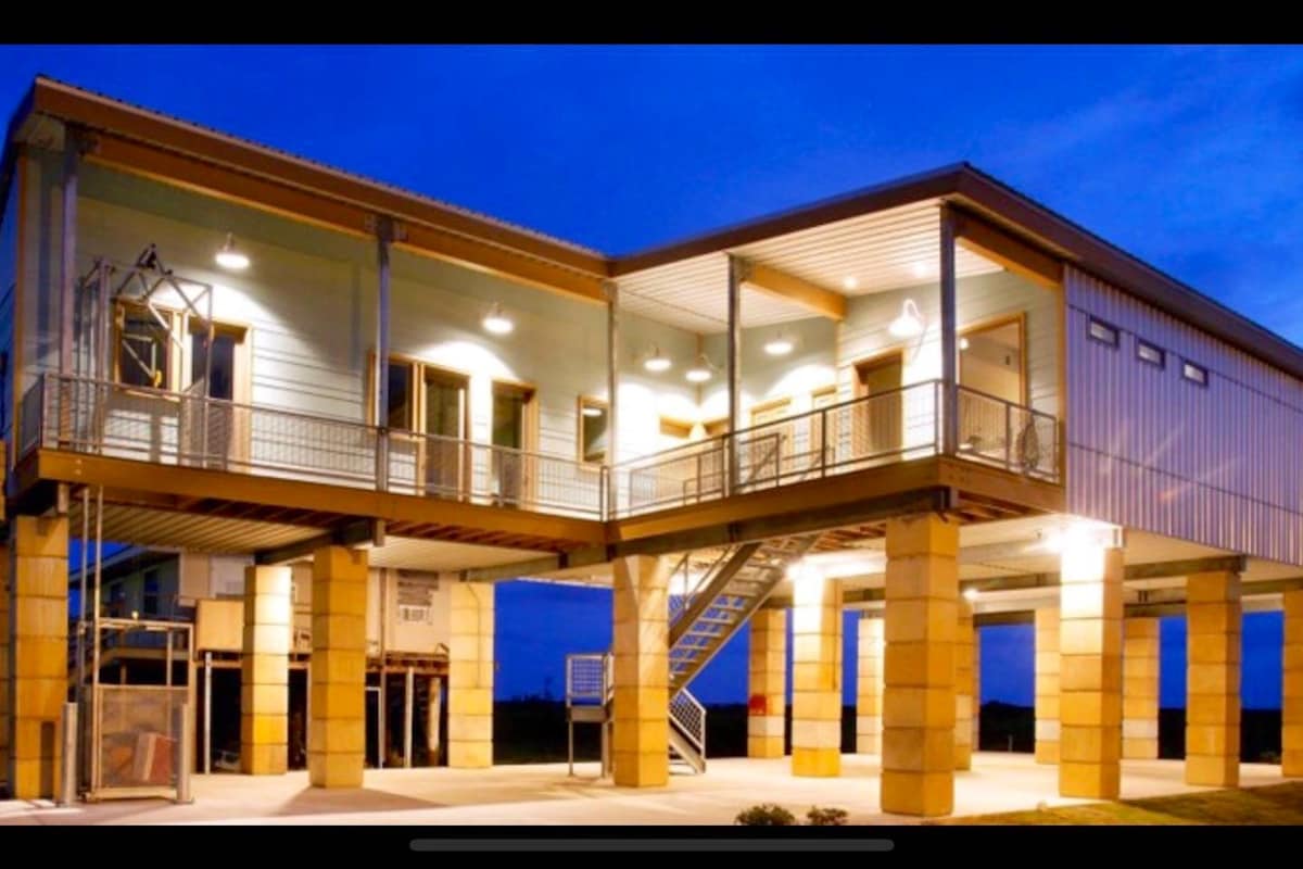 The exterior of a modern two-story building is showcased, featuring a blend of light-colored walls and metal accents. Ground-level support columns elevate the structure, while large windows allow for natural light. Soft illumination from outdoor lights enhances the architectural details against the evening sky.