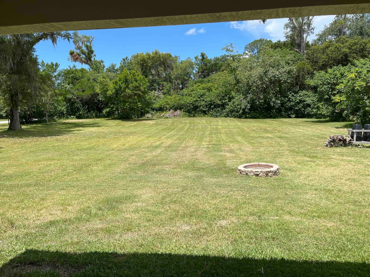 A large, grassy backyard is enclosed by trees and shrubbery, providing a natural, private setting. A stone fire pit is visible in the foreground, complementing the expansive lawn that allows ample space for outdoor activities.
