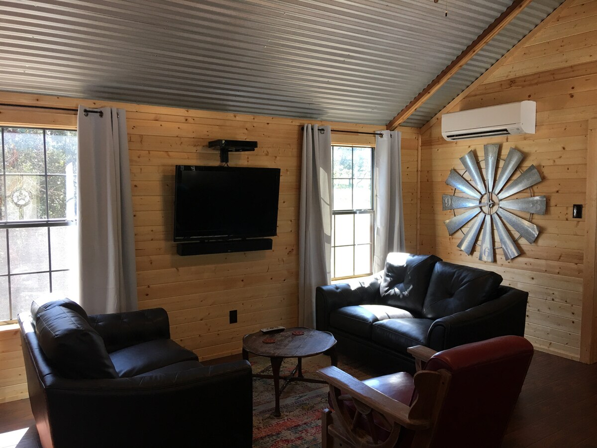 The living area features wooden walls and a corrugated metal ceiling. Two comfortable dark sofas are arranged around a circular coffee table. A flat-screen television is mounted on the wall opposite the seating. Natural light enters through the windows dressed with light-colored curtains.