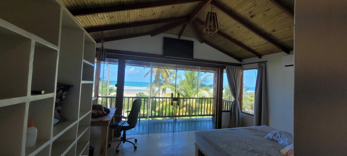 A spacious room is highlighted by a vaulted ceiling and large windows offering ocean views. A desk and chair are positioned near the windows, and a bed is visible with light bedding. Shelves line one wall, providing ample storage and organization.