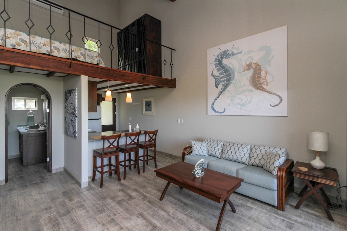 The suite features an open living space with a plush sofa, a wooden coffee table, and a dining area with four wooden chairs. A loft area above contains a king-size bed, visible through a railing. Coastal-themed artwork adorns the wall, adding a touch of serenity.