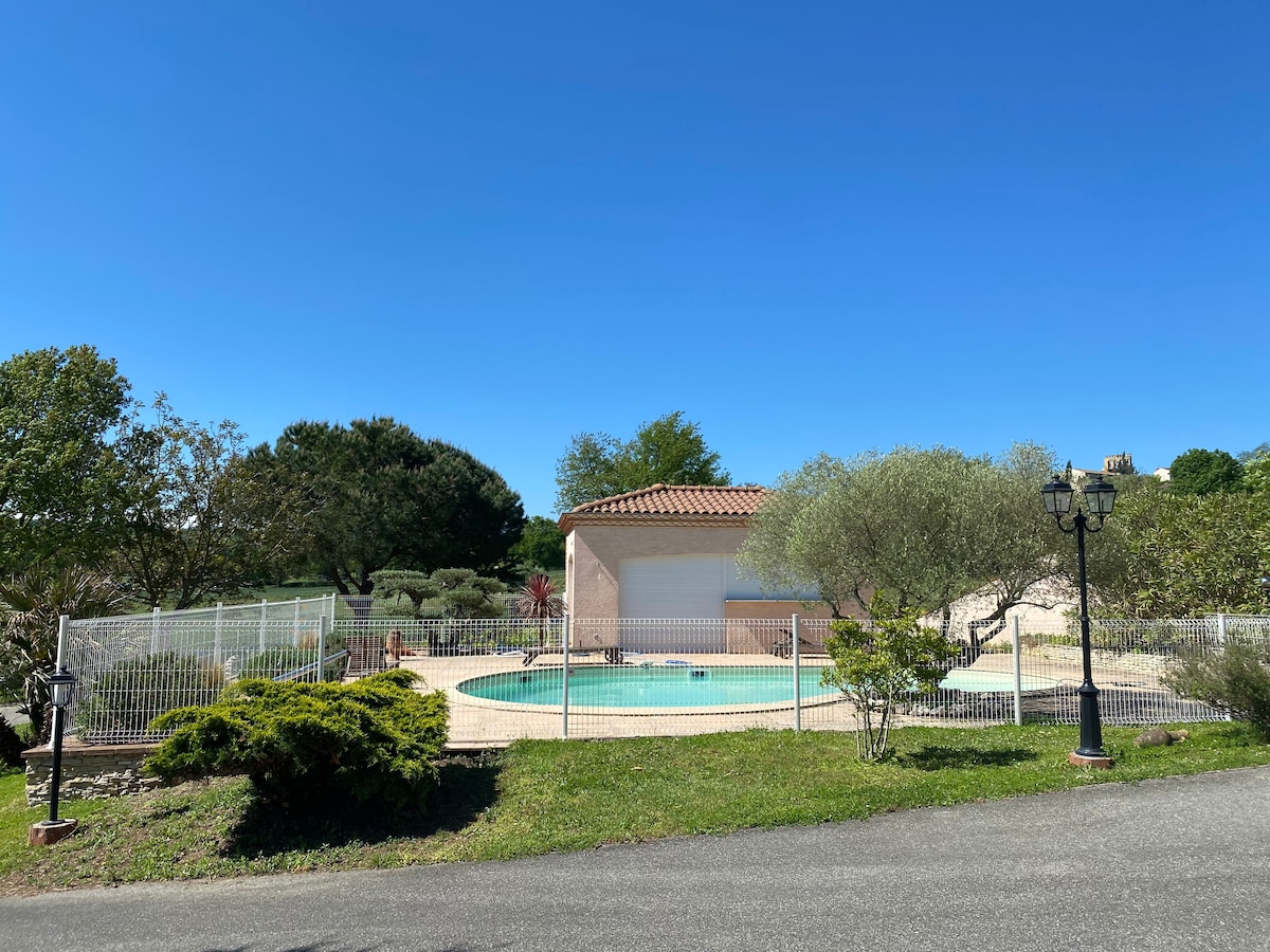 A fenced pool area is surrounded by lush greenery and trees. A light-colored building with a tiled roof is visible in the background. Clear blue skies enhance the inviting outdoor space.