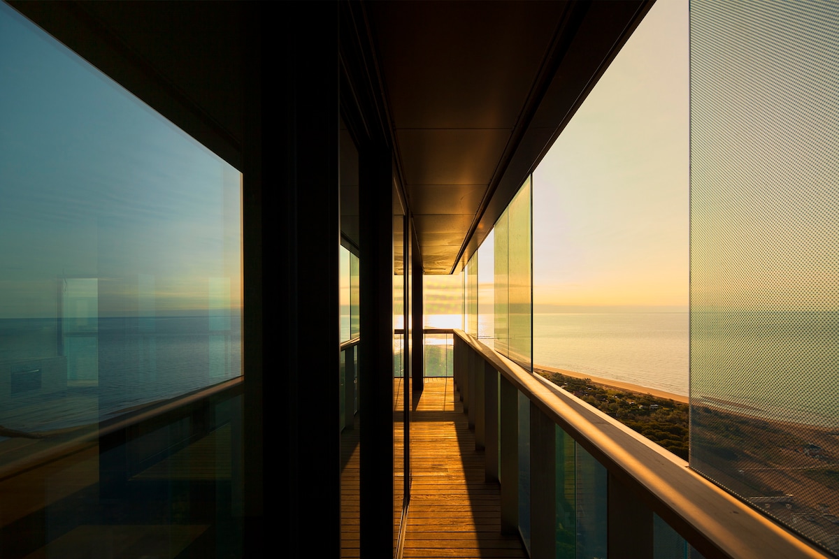 A spacious balcony is depicted, featuring a wooden floor and large glass panels. The sunset reflects off the glass, highlighting views of the sea and coastline. The horizon glows with warm tones, creating a serene atmosphere.