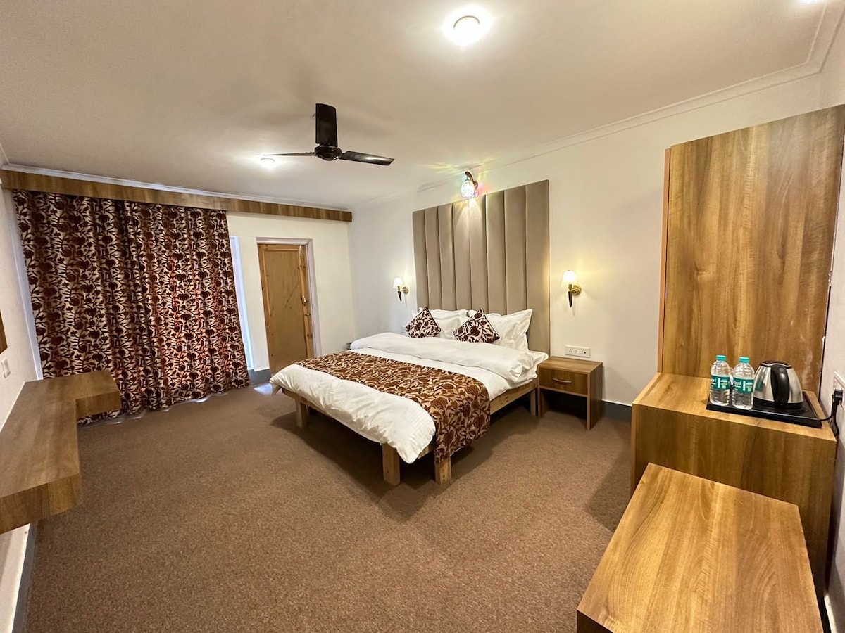 A spacious bedroom features a large bed with a patterned bedspread and matching decorative pillows. Handcrafted crewel curtains frame the window, while a wooden desk and bedside tables complement the warm tones of the decor. An electric kettle and water bottles are provided on the desk.