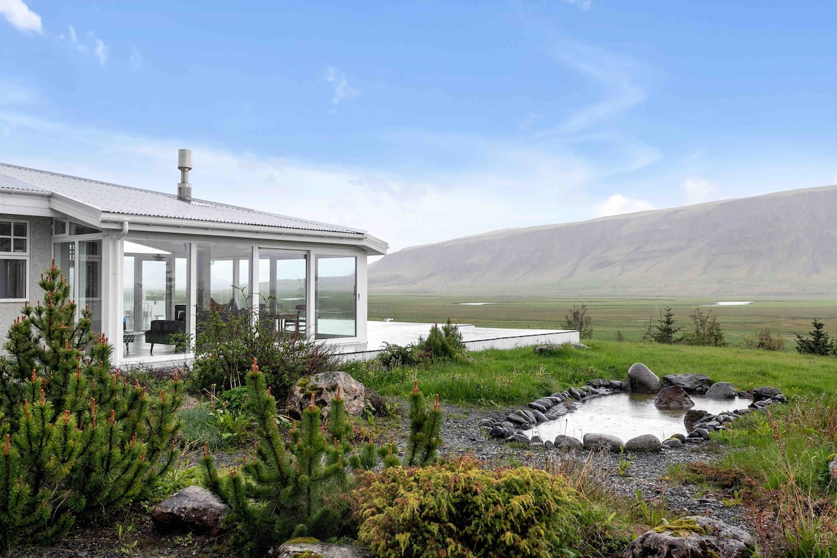 The exterior of the villa presents a modern design with large glass windows reflecting the surrounding landscape. Lush greenery and rocks frame a small natural pond, showcasing the tranquil environment. The expansive view of hills and sky adds to the serene atmosphere.