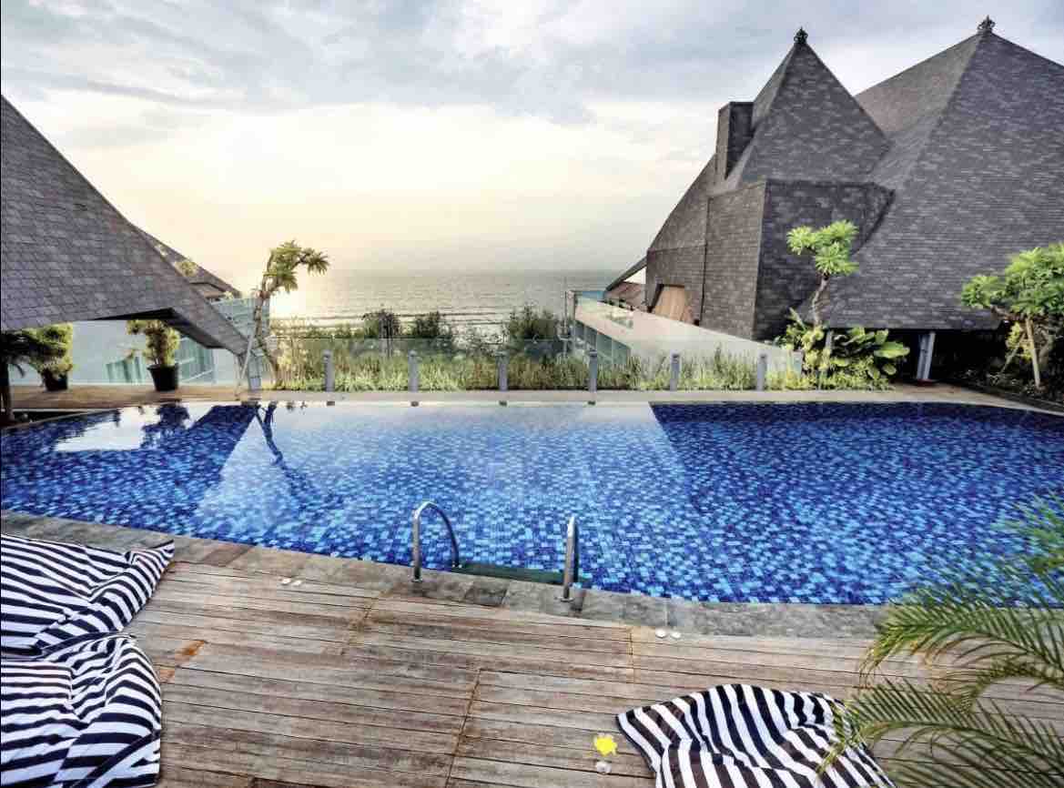 An infinity pool with bright blue tiles extends towards the horizon, reflecting the sky. Surrounding the pool, wooden decking is complemented by striped lounge chairs. Lush greenery adds a natural touch to the modern architectural elements in the background.
