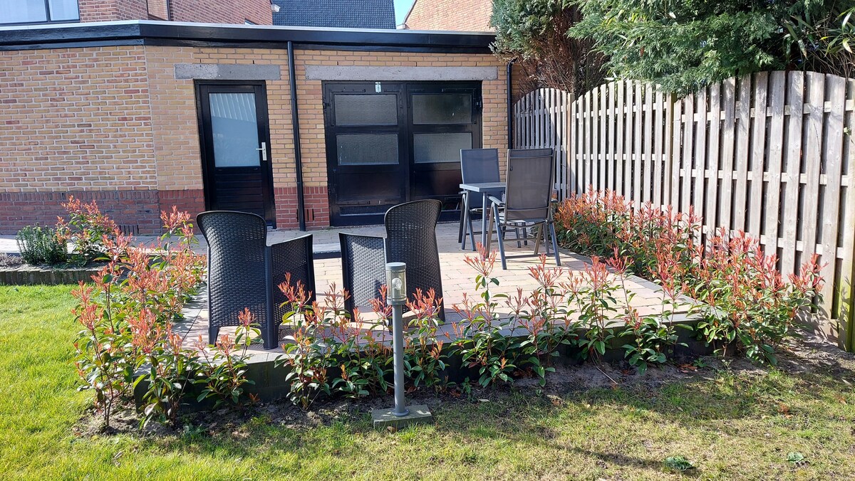An outdoor terrace is furnished with four armchairs, surrounded by blooming greenery. A wooden deck enhances the inviting space, bordered by a wooden fence. The entrance to the accommodation is visible in the background, adding to the private outdoor setting.