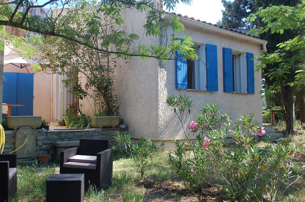 A cozy exterior view of the house reveals a light-colored facade accented by blue shutters. Lush green plants and flowers surround the property, while a shaded seating area is visible, providing a tranquil spot to relax amidst the natural landscape.