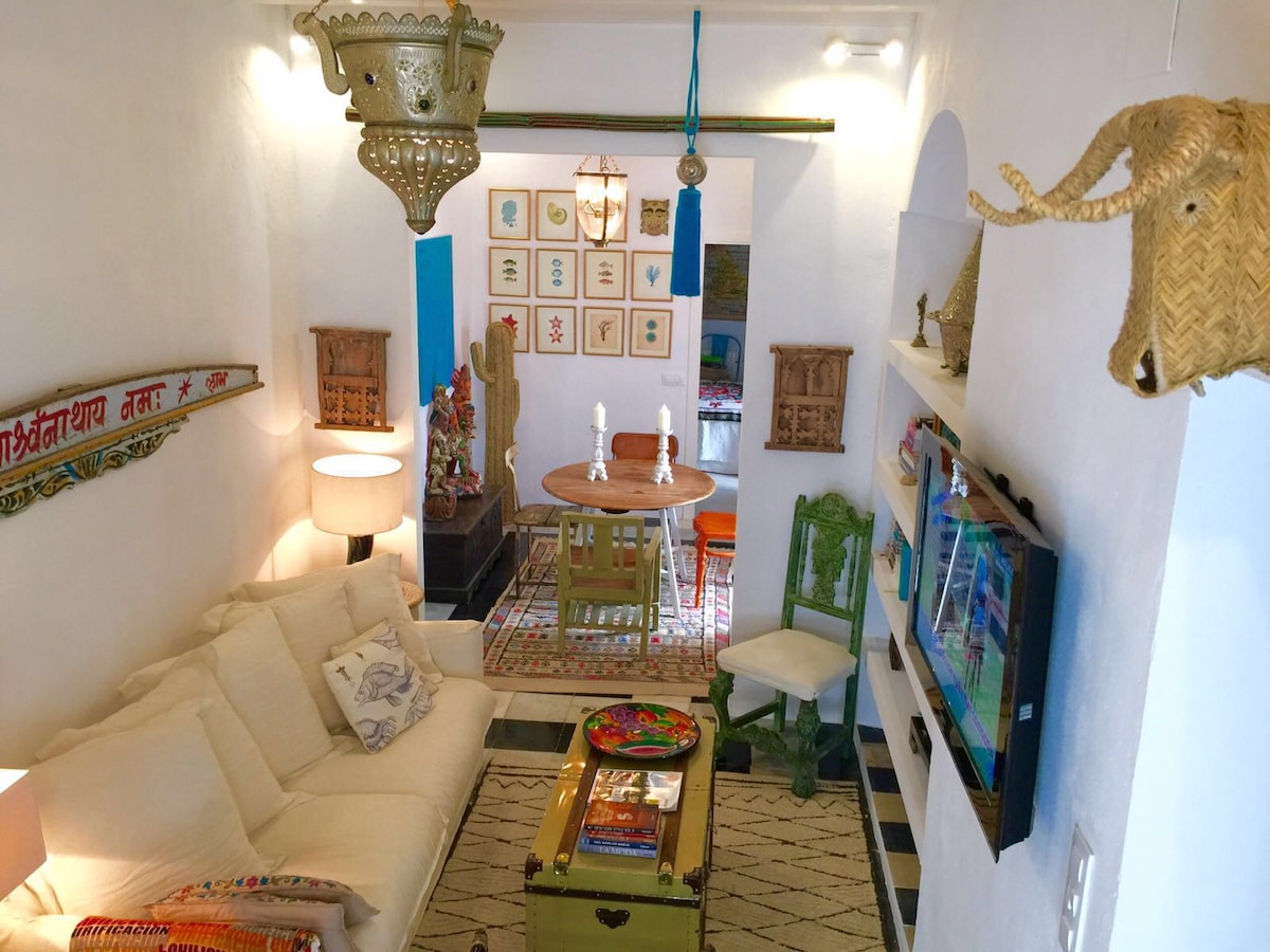 A spacious living area features a light-colored sofa, a round dining table, and decorative elements, including woven wall art and an ornate hanging light fixture. A TV is mounted on the wall. Bright colors and unique decor elements create an inviting space.