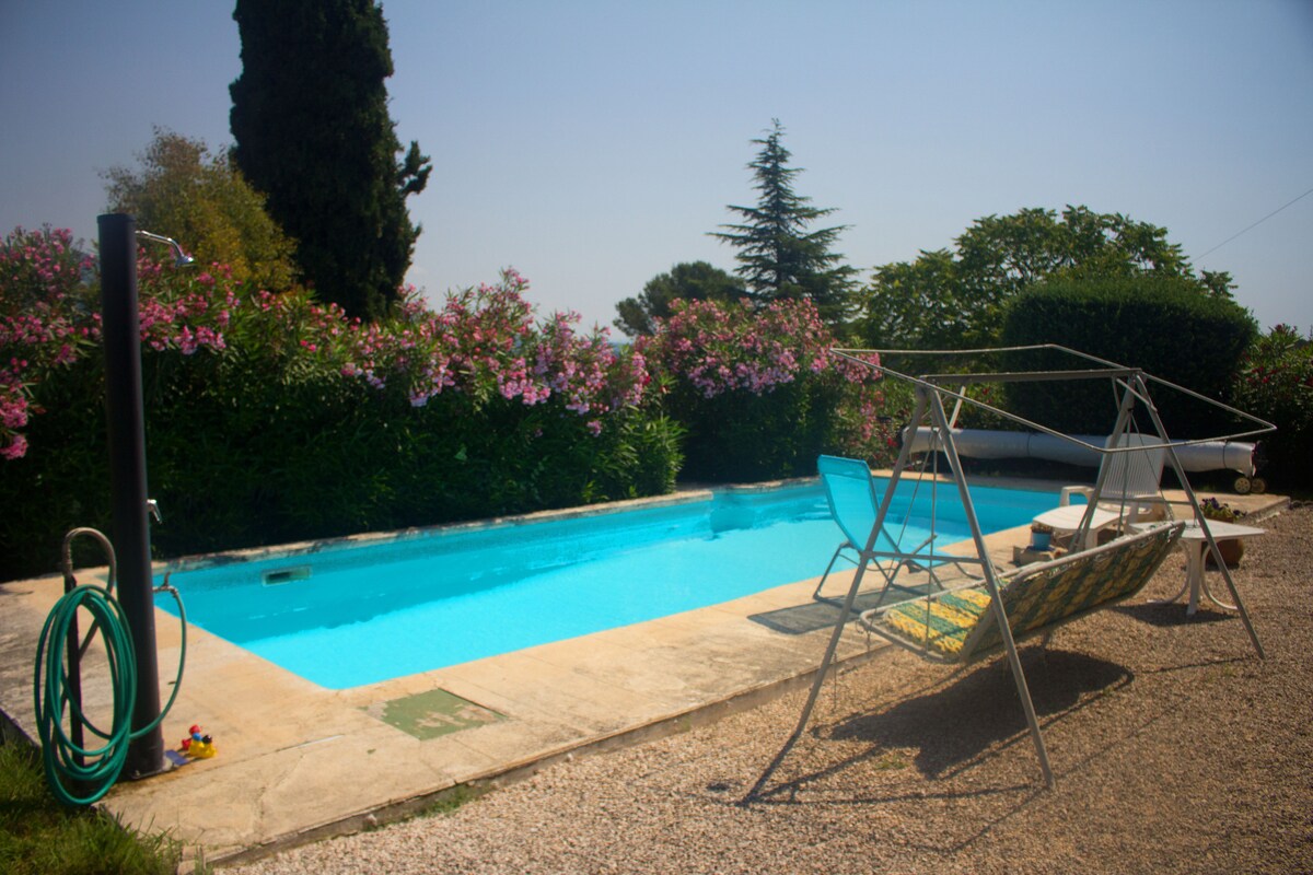 A clear blue pool is surrounded by vibrant flowering plants, enhancing the serene outdoor space. A swing frame is seen beside the pool, inviting relaxation. The charming gravel area provides a comfortable environment for sunbathing or enjoying the view.