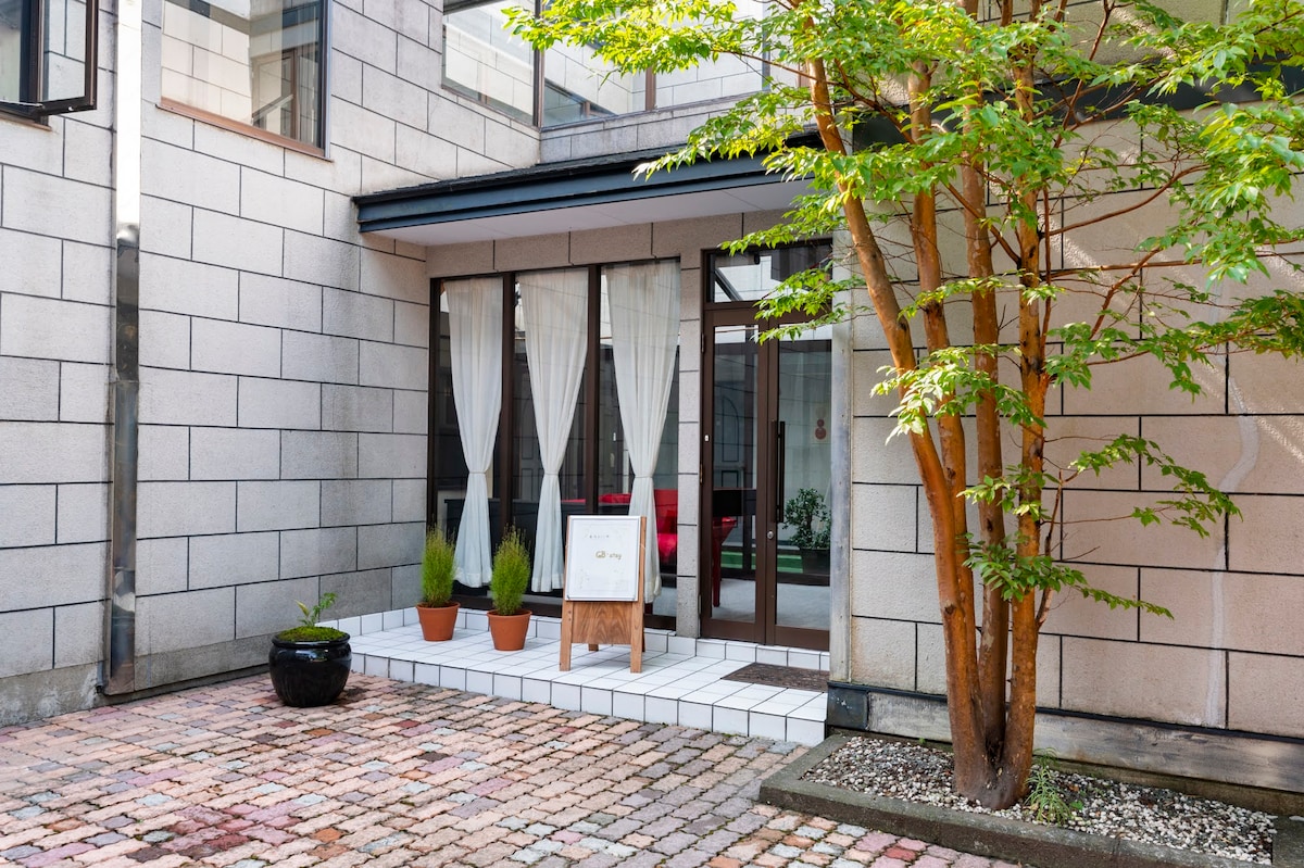 An entrance area is shown, featuring a series of potted plants and a small sign on a stand. The stone pathway leads to a doorway framed by light curtains, surrounded by a shaded patio space with greenery.