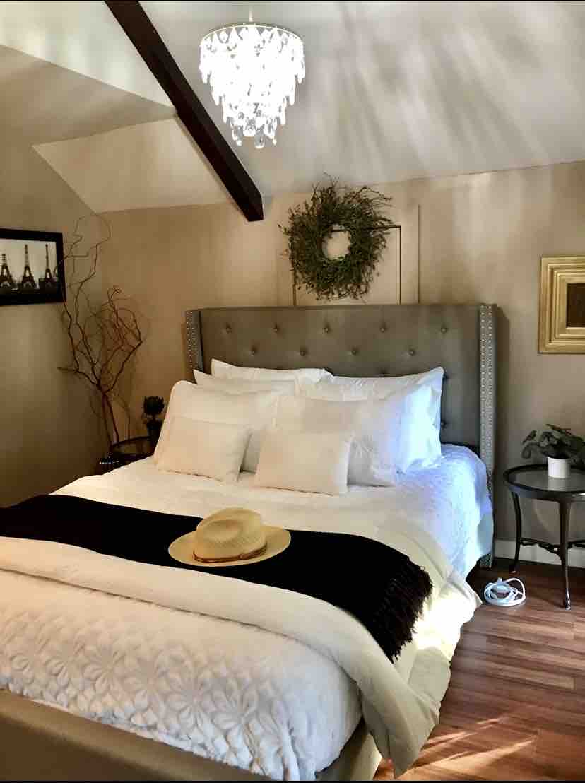 A spacious bedroom features a comfortable bed adorned with multiple pillows and a cozy throw. A stylish chandelier hangs from the ceiling, while natural light filters through nearby windows. Decorative accents include a wall wreath and framed artwork, enhancing the inviting atmosphere.