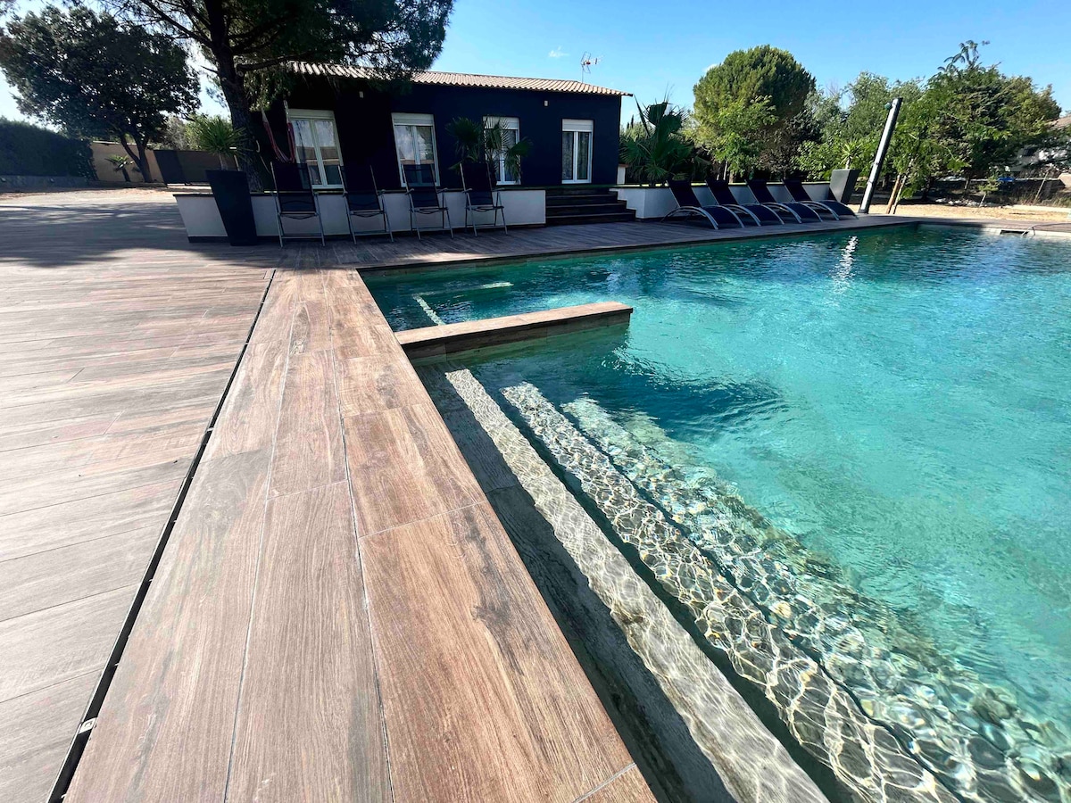 A large swimming pool with a green Bali porcelain finish is surrounded by a wooden deck. Sun loungers are positioned along the edge, and a pathway leads to the entrance of the nearby villa. Lush greenery frames the overall setting.