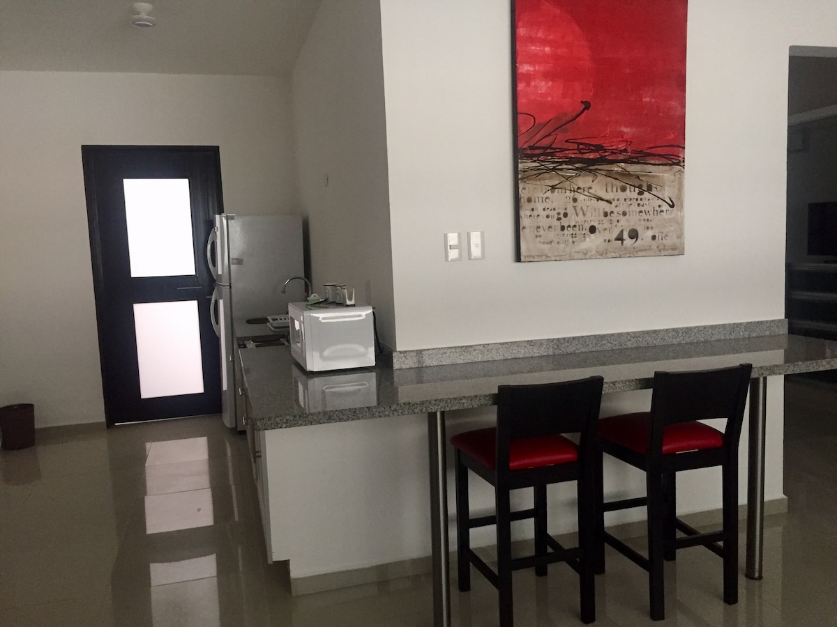 A modern kitchenette features a granite countertop with two stools. A microwave and a refrigerator are visible, alongside a painting with bold red and black accents. Light-colored tiles extend across the floor, enhancing the open atmosphere of the space.