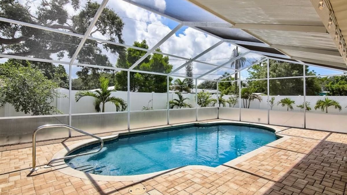 A private heated pool is surrounded by a screened lanai, featuring a subtly shaped design. Lush greenery is visible in the background, providing a natural ambiance. The pool area is paved with light-colored tiles, enhancing the clean and fresh appearance of the space.