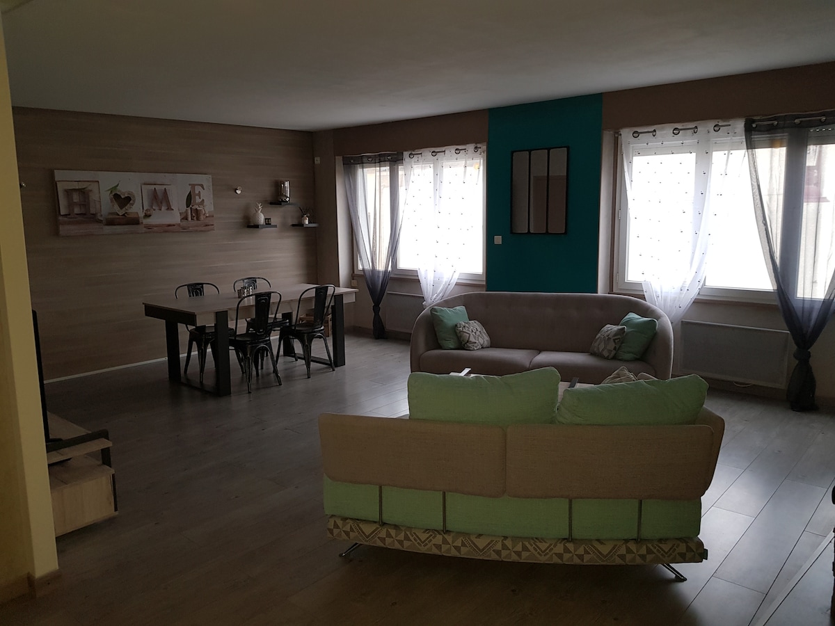 A spacious living area showcases a large, light-colored sofa paired with green and beige cushions. A dining table with several chairs is positioned nearby, and large windows allow natural light to fill the room, which features a calming color palette and decorative wall displays.