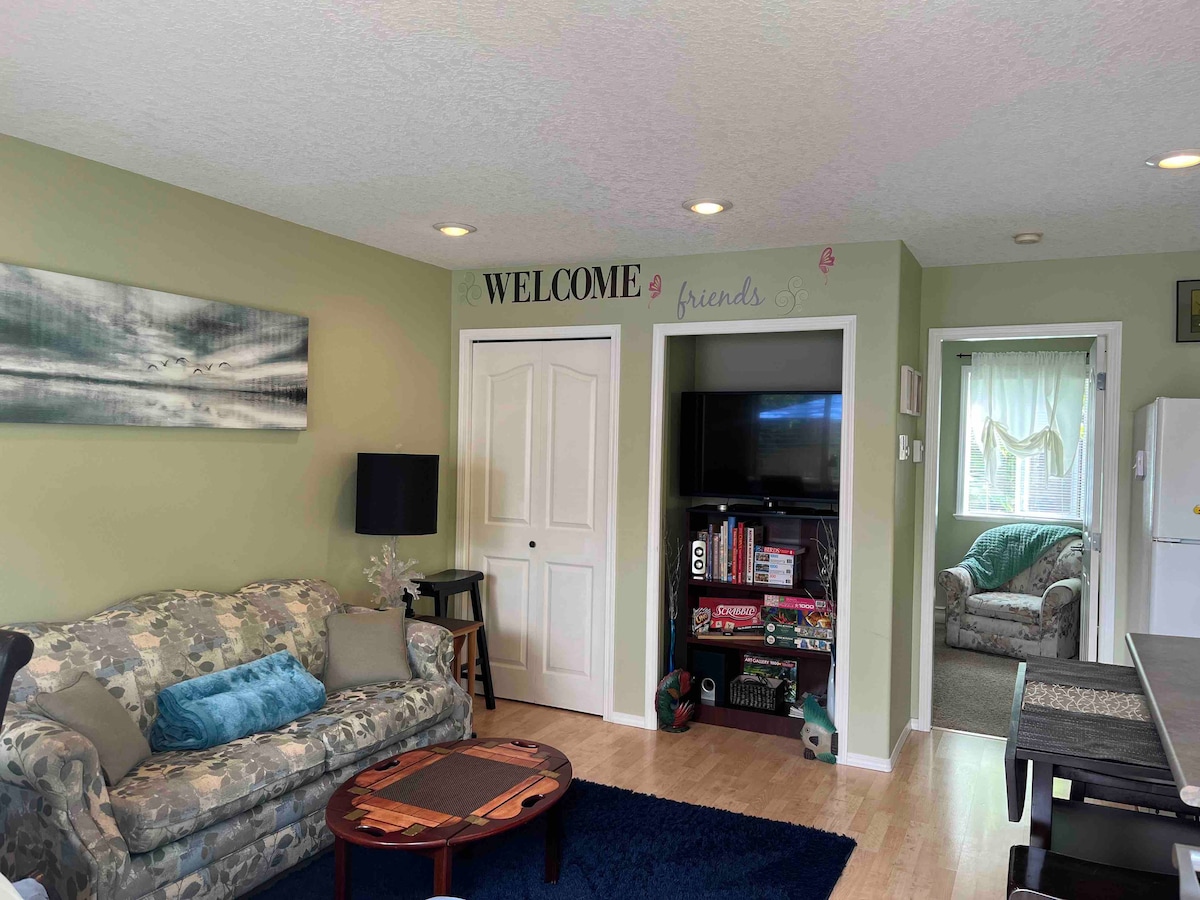 A cozy living area is highlighted by a camouflaged-patterned couch adorned with decorative pillows. A circular coffee table sits in front, while a television is positioned within a built-in shelf. Natural light filters through a window, illuminating the welcoming atmosphere.