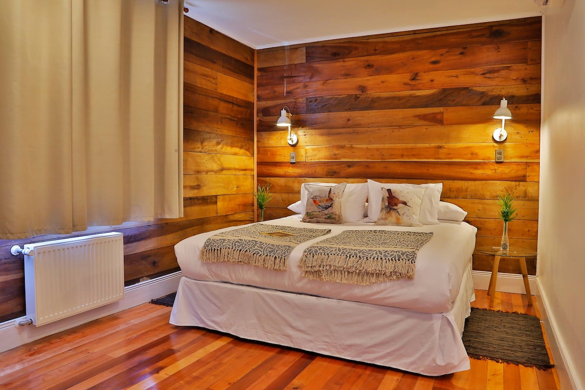 A cozy sleeping area features a spacious bed adorned with decorative pillows and a patterned throw blanket. Warm wooden paneling lines the walls, complemented by soft lighting from mounted fixtures. A radiator and small plants add functional touches to the inviting space.