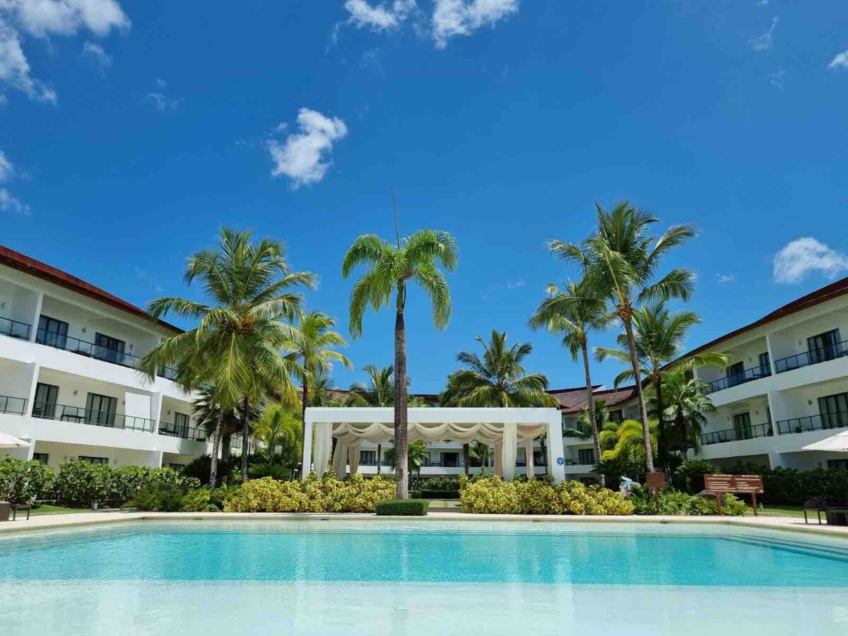 A pristine swimming pool is framed by lush greenery and palm trees, reflecting a clear blue sky. The elegant architecture of the hotel buildings can be seen in the background, complemented by shaded lounge areas and vibrant landscaping.
