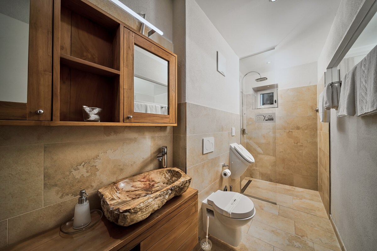The bathroom features natural stone tiles in warm tones. A wooden vanity with a unique basin is complemented by a wall mirror. A modern shower and toilet are present, along with neatly arranged towels on a bar. Soft lighting enhances the serene atmosphere.