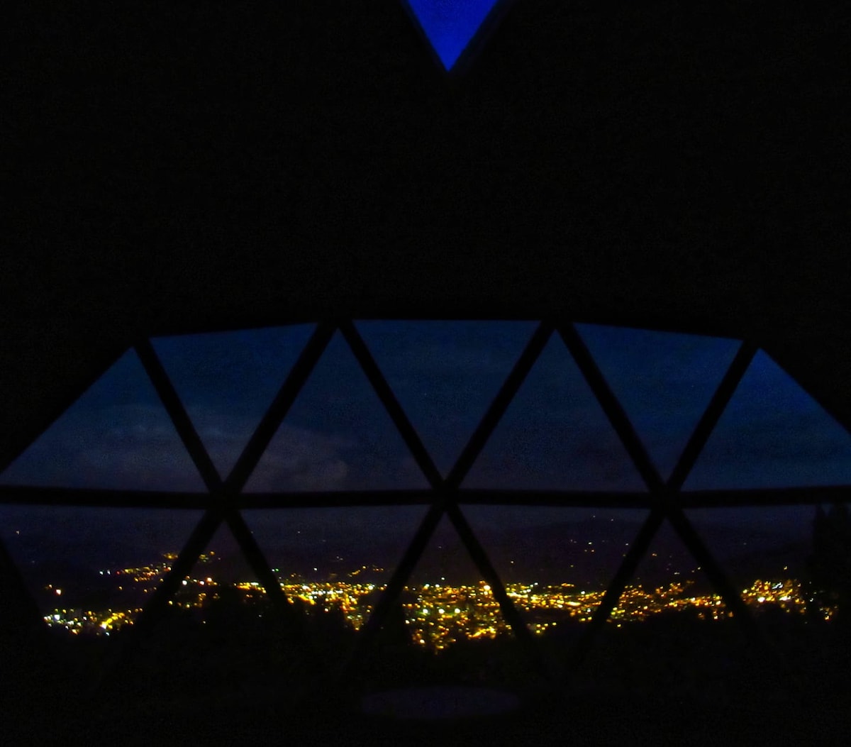 A panoramic view is framed by triangular windows, showcasing the illuminated city of Duitama against a night sky. The gentle glow from the city lights contrasts with the darkness outside, creating a serene atmosphere inside the wooden dome.