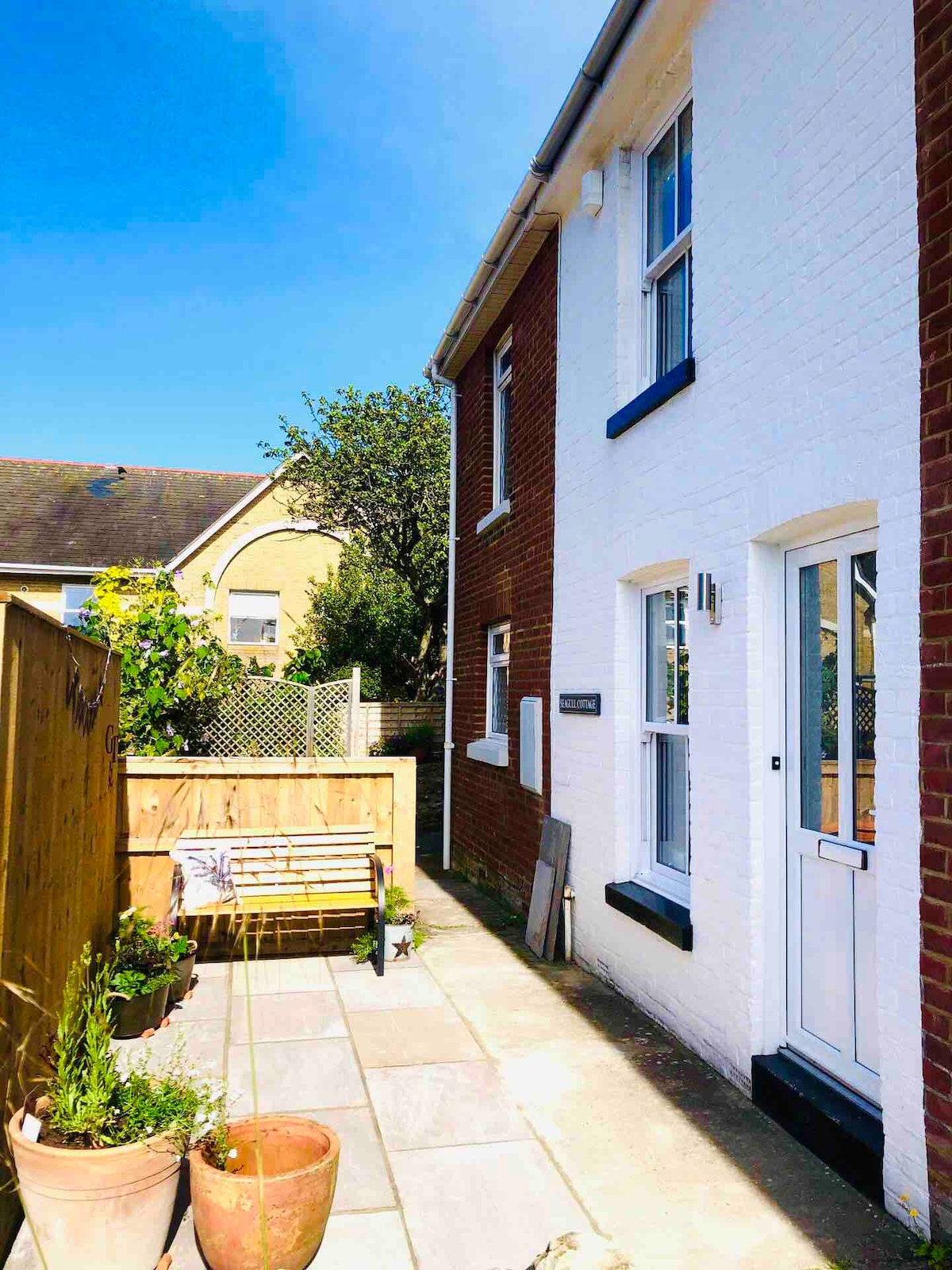 A charming exterior view of Seagull Cottage is presented, featuring a well-maintained pathway leading to the front door. Potted plants and a wooden bench are set against a backdrop of white and red brick walls, surrounded by sunny outdoor spaces with a clear blue sky above.