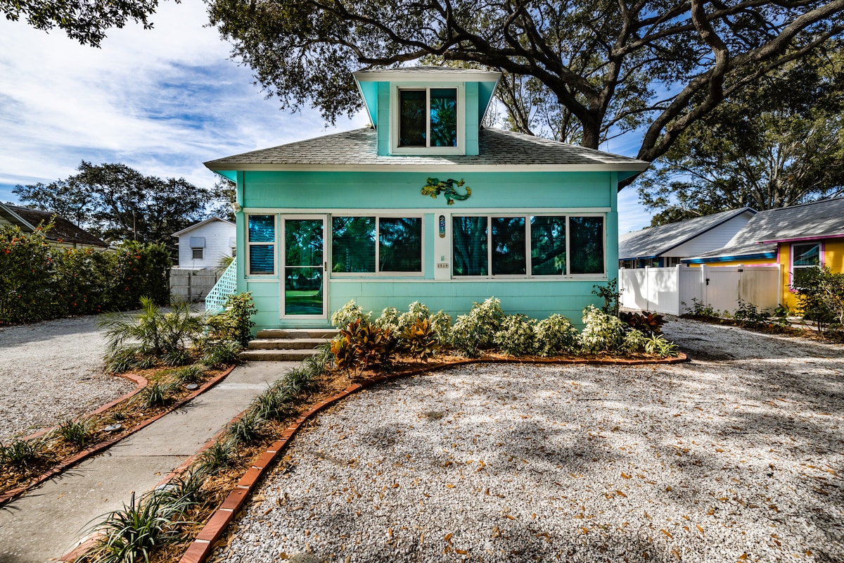 A charming two-story house is presented in soft turquoise, featuring large front windows that invite natural light. A gravel pathway leads to the entrance, bordered by lush greenery and decorative landscaping. The surrounding area includes trees providing shade and a sense of tranquility.