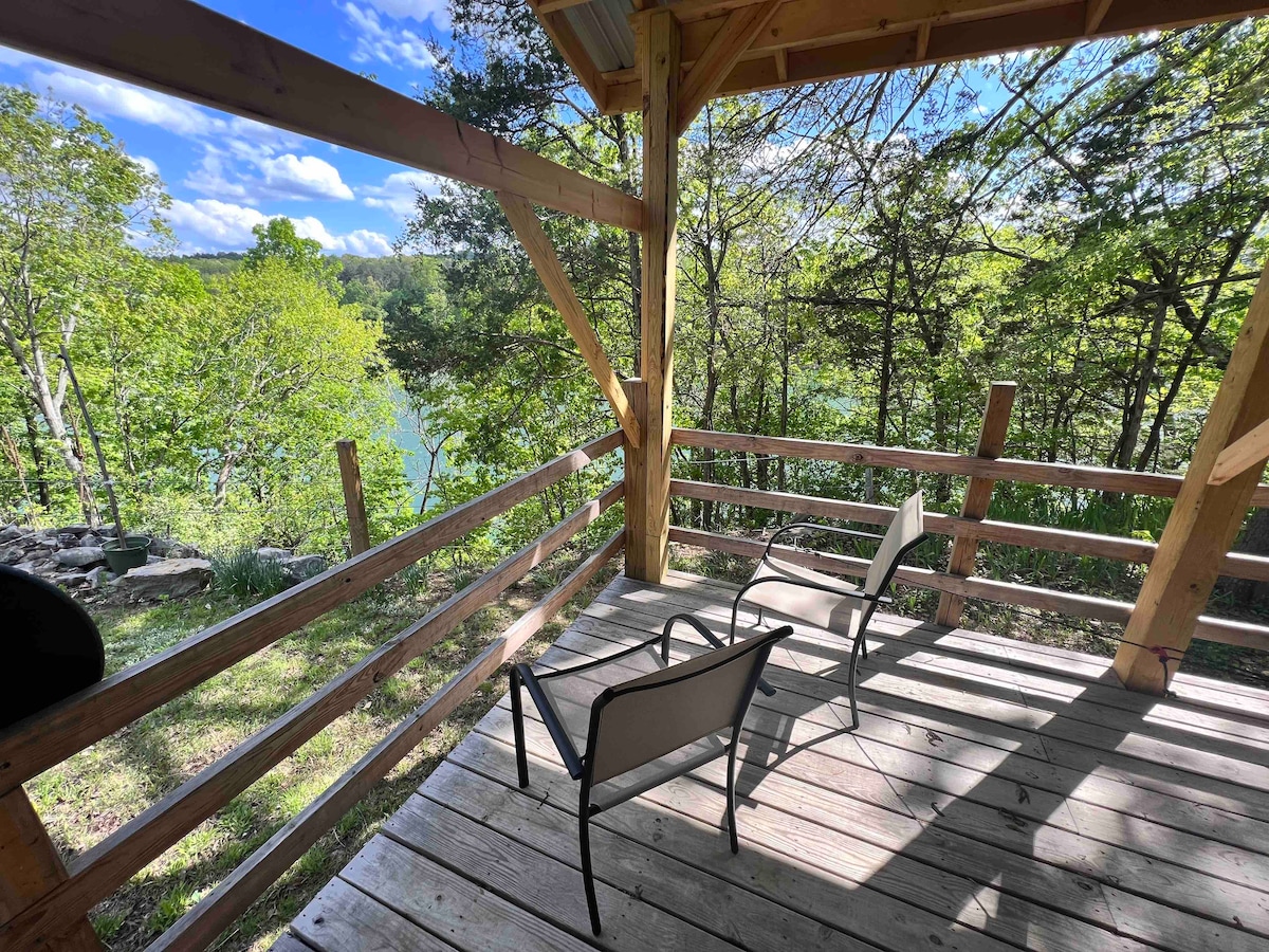 A covered wooden deck features two seating chairs positioned overlooking a lush green landscape. The clear lake is visible through trees in the background, with a mix of sunshine and shadow casting a calming ambiance on the space.