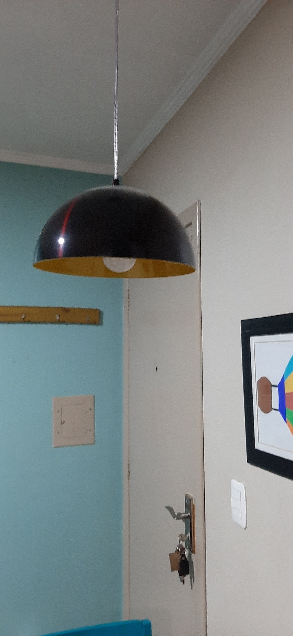 A contemporary pendant lamp hangs from the ceiling, featuring a rounded black exterior with a contrasting yellow interior. The lamp illuminates the space, while a light switch and door handle are visible on the adjacent wall, alongside a colorful geometric artwork.