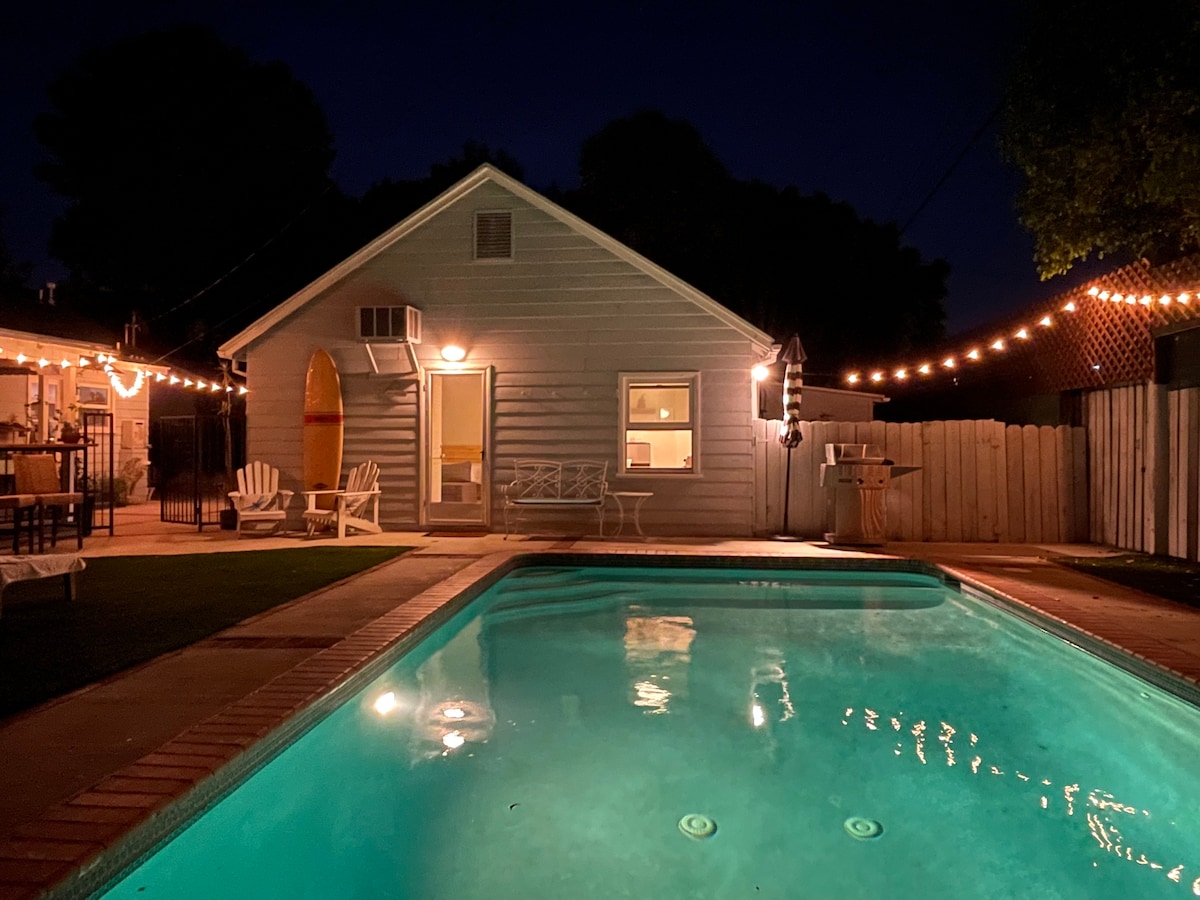 A calming nighttime view of the guest house illuminated by warm string lights. The salt-water pool reflects soft light, enhancing the tranquil setting. Adjoining lounge chairs and a grilling area are seen in the background, creating an inviting outdoor space.