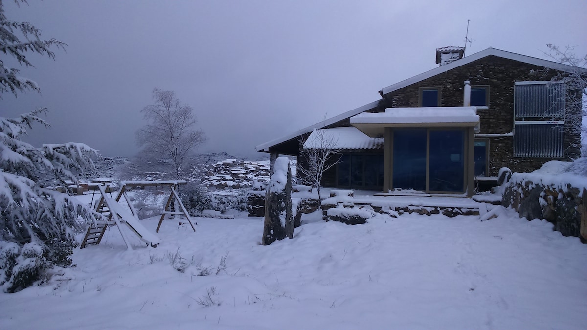 A rustic house is set against a snowy landscape, featuring large glass windows that reflect the surrounding nature. A wooden swing set is visible in the foreground, surrounded by a blanket of snow, while snow-covered trees frame the scene.