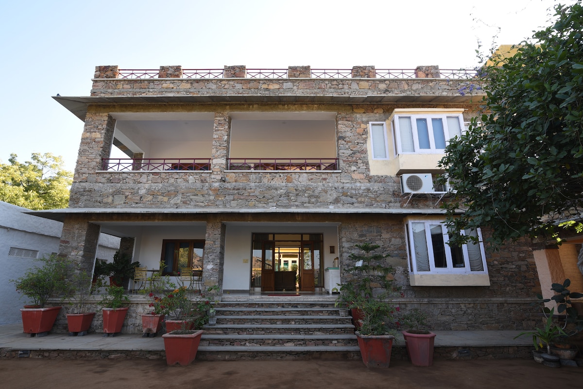The stone exterior of the building showcases a robust design, with a spacious entrance framed by potted plants. The upper balcony features a railing, and large windows allow natural light to flood the interior. Stairs lead up to the entrance, enhancing accessibility.