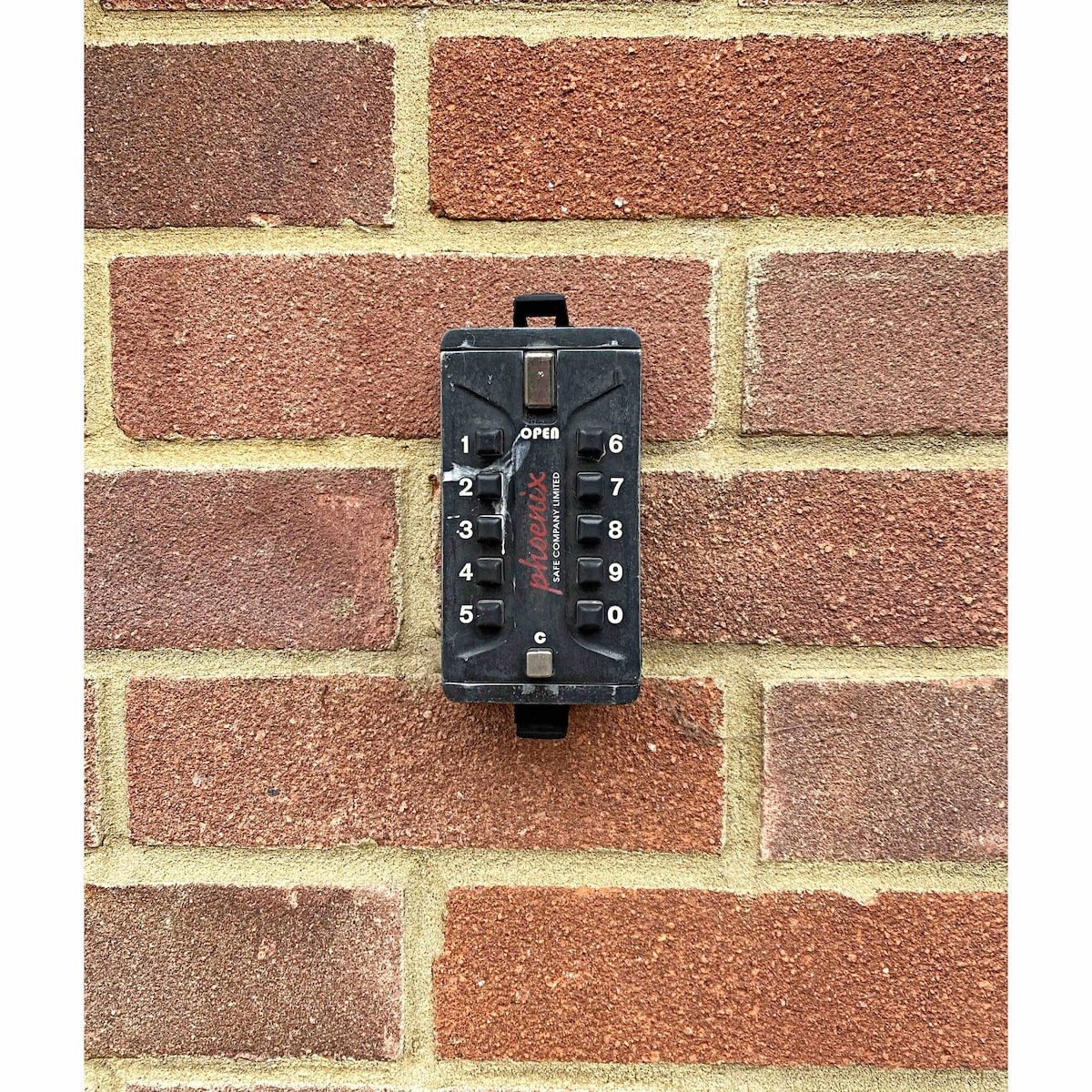 An electric key safe is mounted on a brick wall, featuring a numbered dial for code entry. The black device contrasts with the red and brown tones of the surrounding bricks, providing a secure access point for guests.