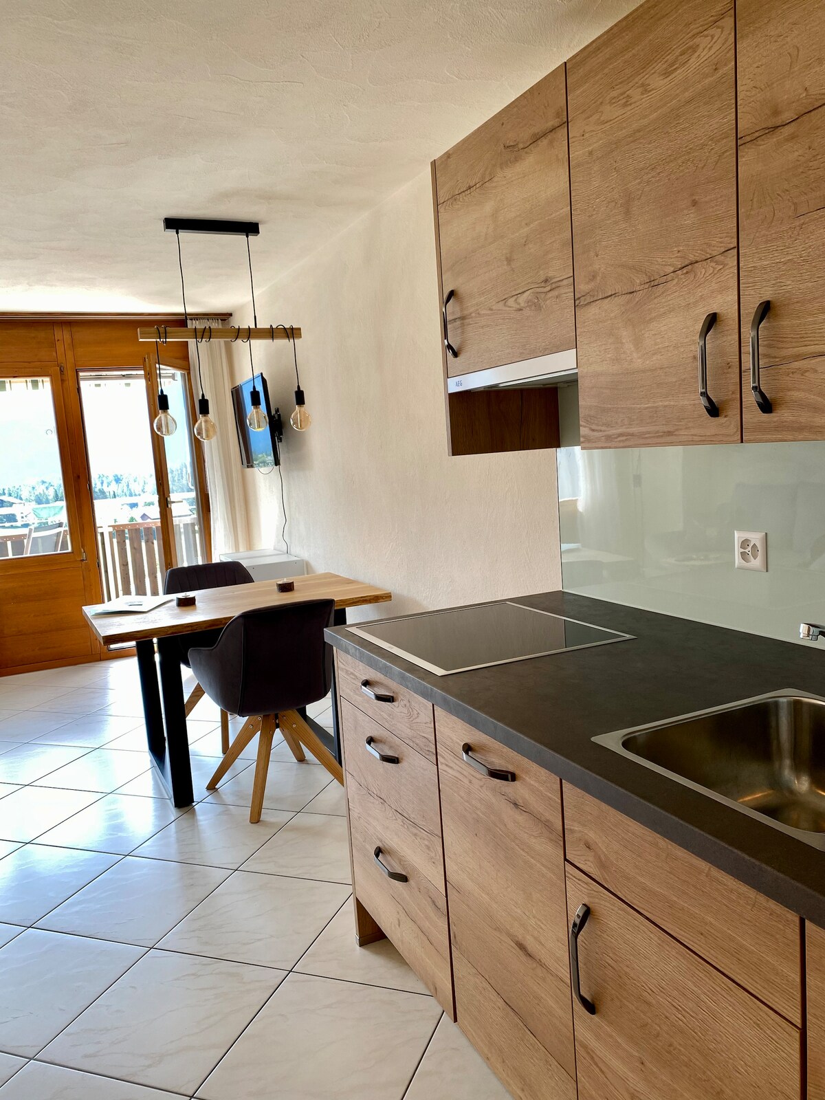 A modern kitchen features wooden cabinetry and a sleek countertop. An induction cooktop is installed, alongside a stainless steel sink. A dining table with Scandinavian-style chairs is positioned near the window, allowing for natural light and scenic views of the surrounding area.