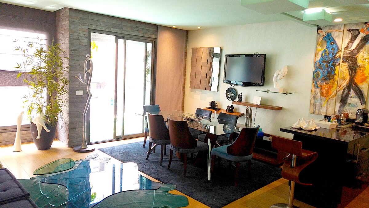 The living and dining area features large glass doors allowing natural light. A contemporary artwork adds color to the space, complemented by elegant furnishings, including a glass dining table and four upholstered chairs. A decorative sculpture and a unique area rug enhance the room's character.