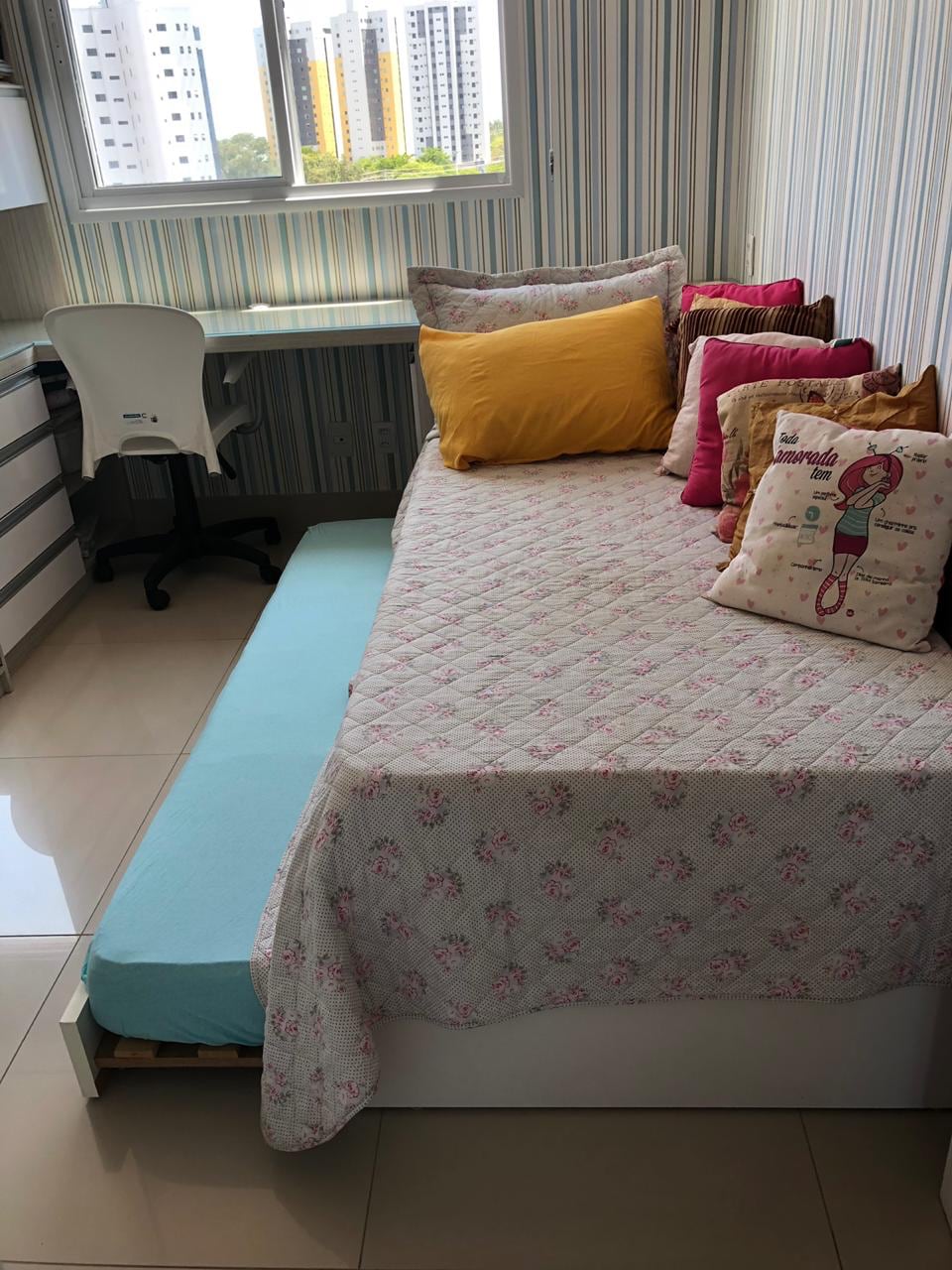 A well-lit room features a daybed adorned with various colorful pillows. A light blue mattress extends from under the bed. A desk with a chair is positioned nearby, and a window allows natural light to brighten the space.