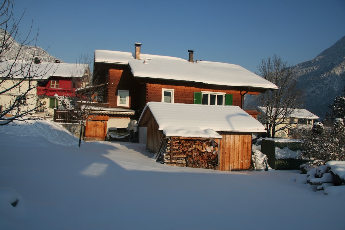 The chalet is surrounded by a blanket of snow, with a wooden shed and stacked firewood visible in the foreground. The sloped roof is covered in snow, and the structure features large windows and a vibrant green window frame against a clear blue sky.