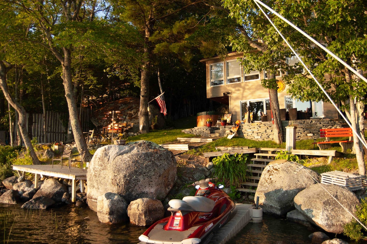 A lakeside house sits amid lush greenery, featuring large windows that invite natural light. A wooden dock extends into the water, with a jet ski parked beside large rocks. An American flag flutters gently in the breeze, enhancing the natural setting.
