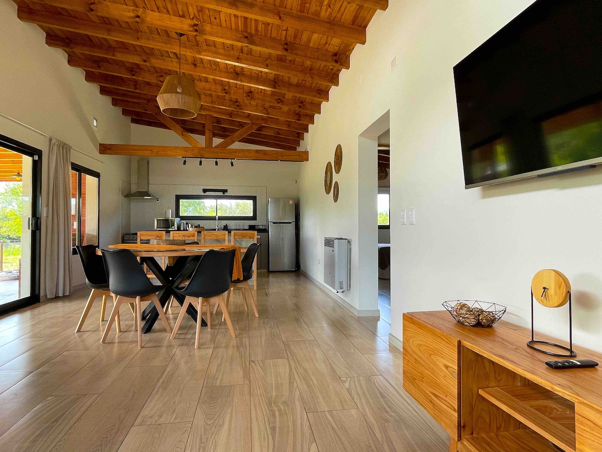 An open-concept living and dining area features a wooden ceiling with exposed beams. A dining table with black chairs is centrally located, complemented by a wooden sideboard and wall-mounted television. Expansive glass doors lead to the outdoor space, providing abundant natural light.
