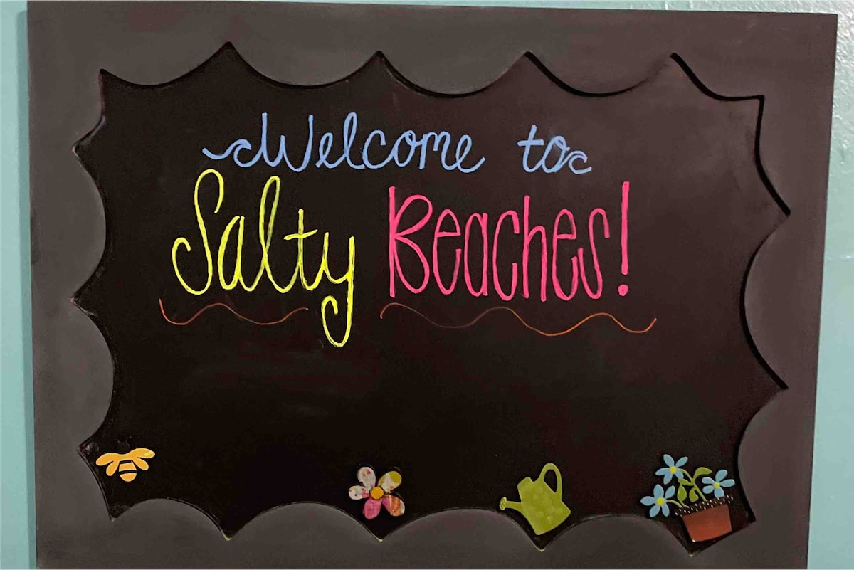 A decorative sign displayed on a wall, featuring the text 'Welcome to Salty Beaches!' written in colorful chalk. Floral and garden-themed illustrations adorn the corners, adding a cheerful touch.