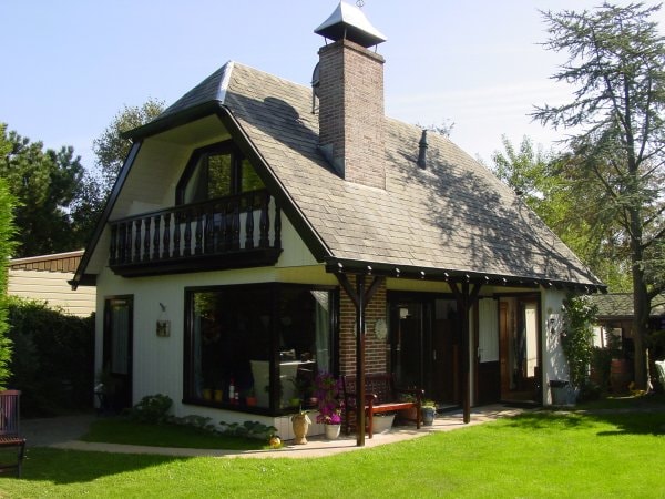 A charming two-story house with a sloped roof and a prominent chimney is surrounded by lush greenery. Large windows allow natural light to fill the interior, while a balcony above adds an inviting touch. The well-maintained garden provides a serene outdoor space.