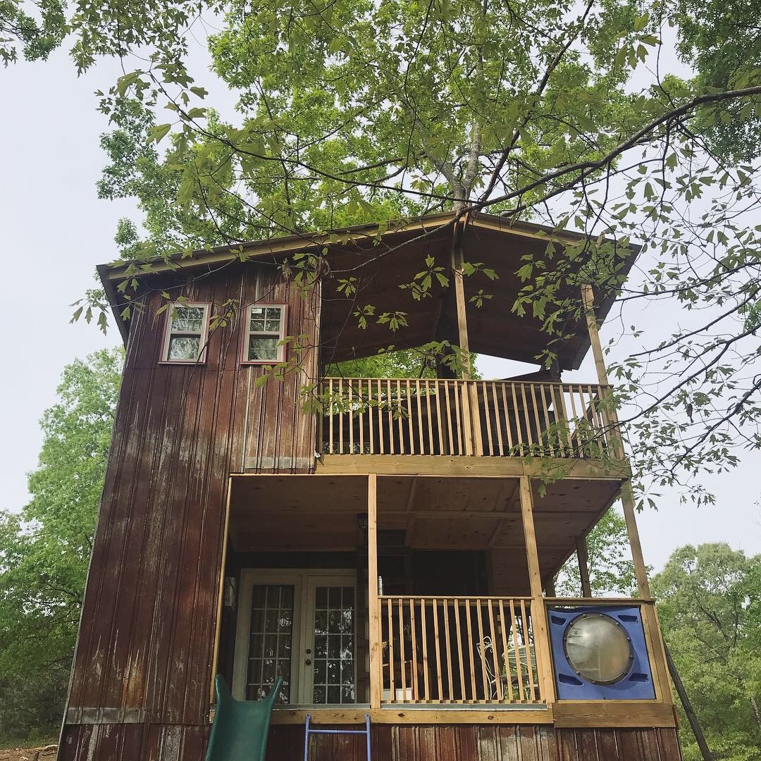A two-story wooden cabin is embraced by greenery, featuring a rustic exterior with red-stained planks and a sloped roof. A spacious balcony is visible on the upper level, with wooden railings and a slide attached on the side, inviting playful exploration.
