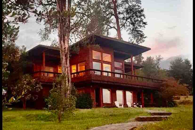 A two-story wooden house features large windows that reflect warm light, surrounded by lush greenery. A spacious terrace extends around the building, providing a serene outdoor space. Pathways lead through the garden, enhancing accessibility to the natural surroundings.