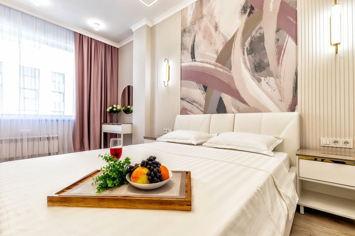 A well-appointed bedroom features a spacious bed with white linens. A decorative tray with fruit and a beverage resides on the bed. Soft curtains frame a large window that allows natural light to fill the space, while a stylish wall mural complements the decor.